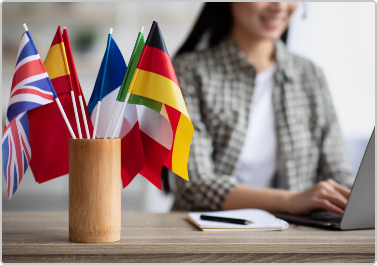 mini flags of European countries in a wooden cup in foreground of woman on a laptop in background